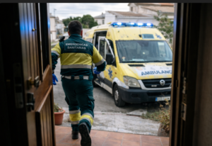 Técnico en emergencias sanitarias de espaldas con uniforme verde y amarillo saliendo por una puerta hacia una ambulancia amarilla
