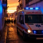 Ambulancia con las luces de emergencia encendidas ante una calle cortada por varias personas al atardecer en un barrio andaluz